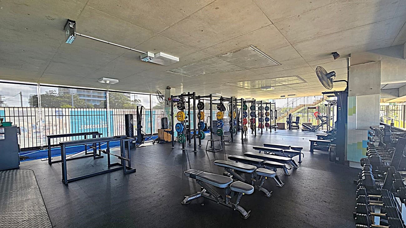 A spacious gym with a black rubber floor and a concrete ceiling. On the left, there are weightlifting platforms and racks with colourful weight plates. In the centre, several adjustable benches are lined up. On the right, a row of dumbbells is neatly arranged on racks. The background features large windows with metal bars, allowing natural light to illuminate the space. Ceiling fans and lights are visible above.