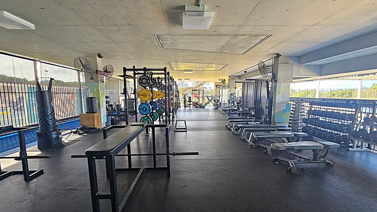 A spacious gym with a variety of equipment. On the left, there are punching bags and a rack of weight plates in various colours. In the centre, there are benches and a squat rack. On the right, there are several benches and a rack of dumbbells. The floor is covered with black rubber mats, and the ceiling is plain with fluorescent lights. Large windows line the walls, allowing natural light to fill the space.