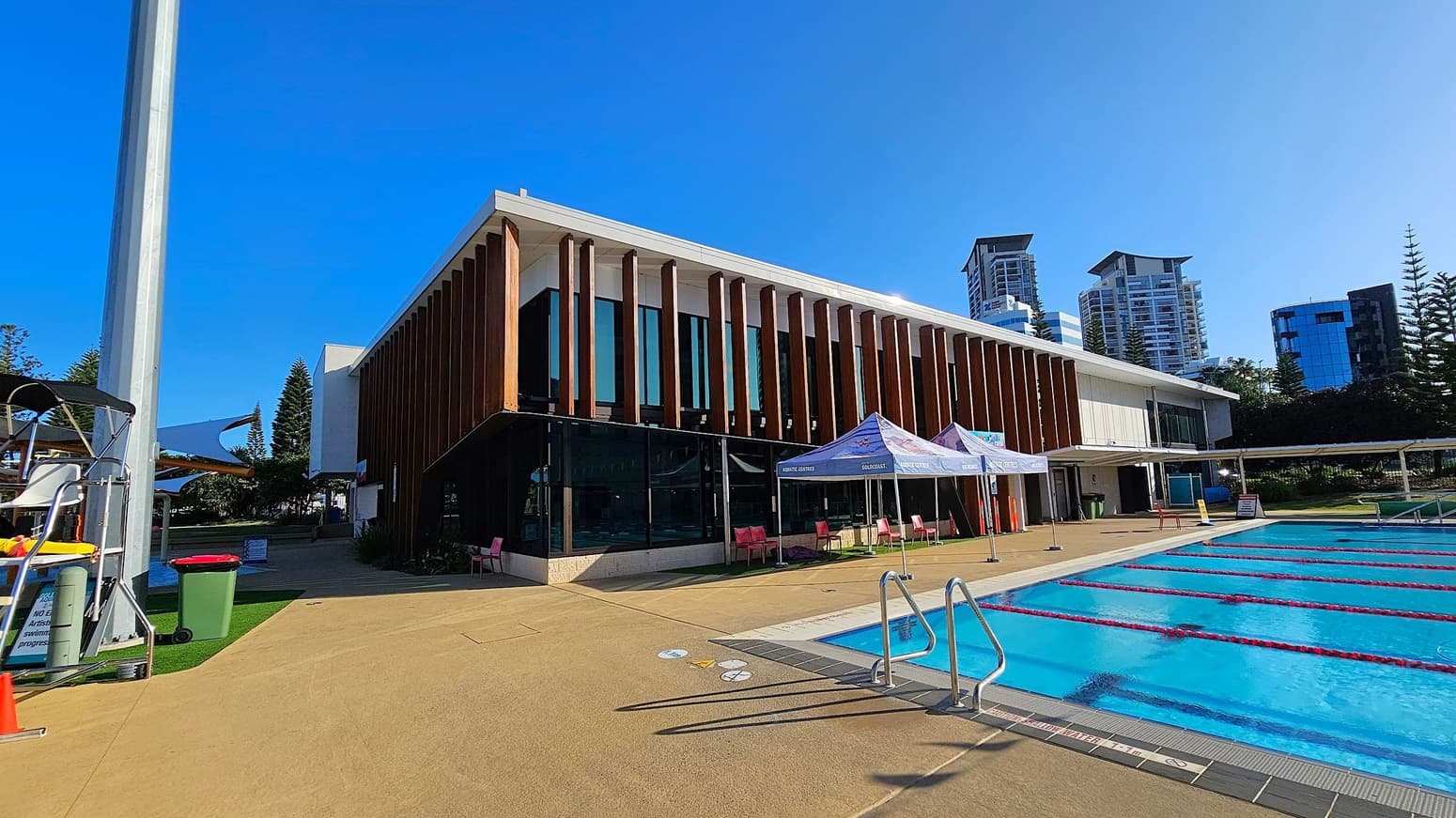 A modern building with vertical wooden slats and large glass windows is on the left. In front, there are two tents with signage, and several red chairs are scattered around. A swimming pool with lane dividers is on the right, bordered by a beige deck. In the background, tall buildings and trees are visible. A tall pole and a green bin are on the left side of the image.