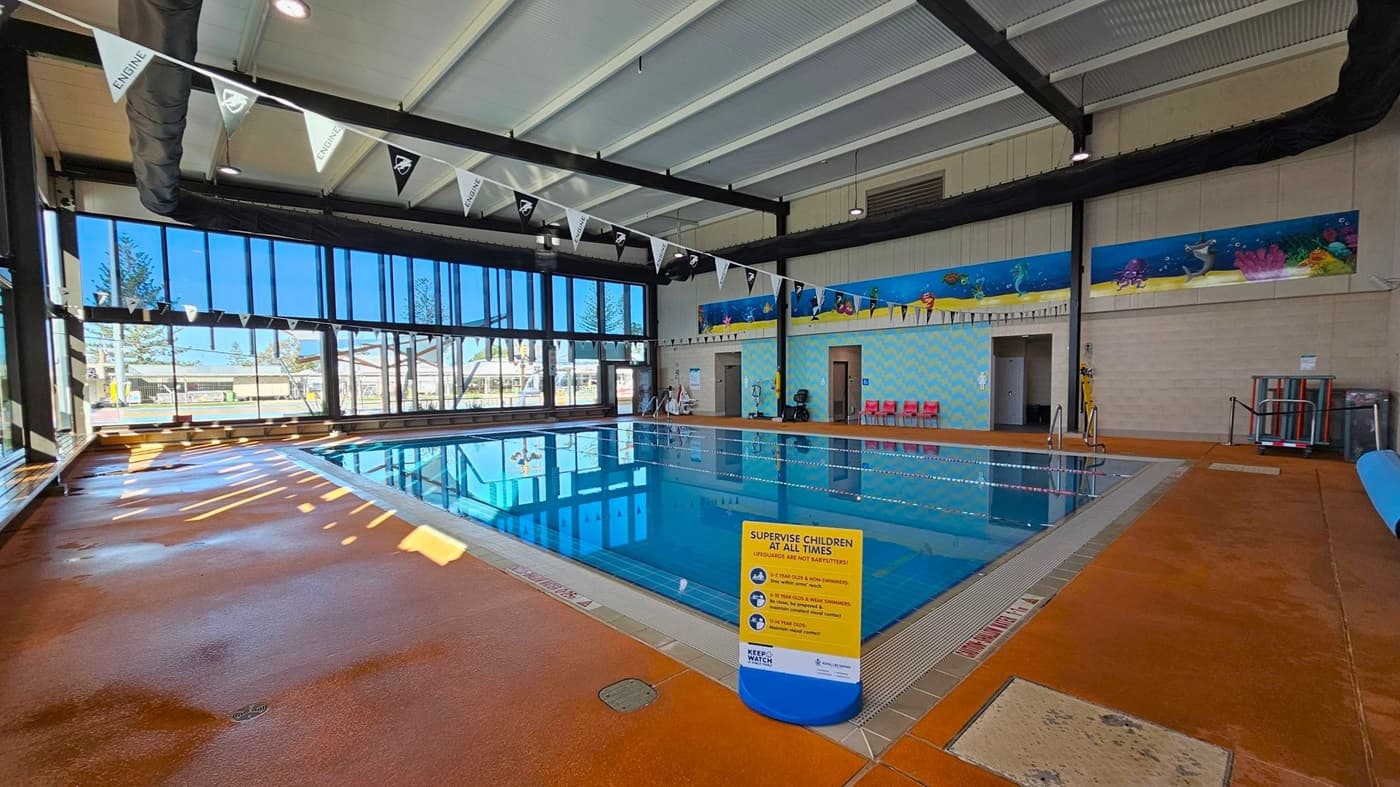 Indoor swimming pool with clear blue water, surrounded by a wet, orange-tiled floor. On the left, large glass windows allow natural light in, with a view of trees outside. A row of triangular flags hangs above the pool. On the right, a colourful mural with sea creatures decorates the wall, above a row of red chairs. A sign near the pool edge provides safety instructions. The ceiling is high with exposed beams and ductwork.