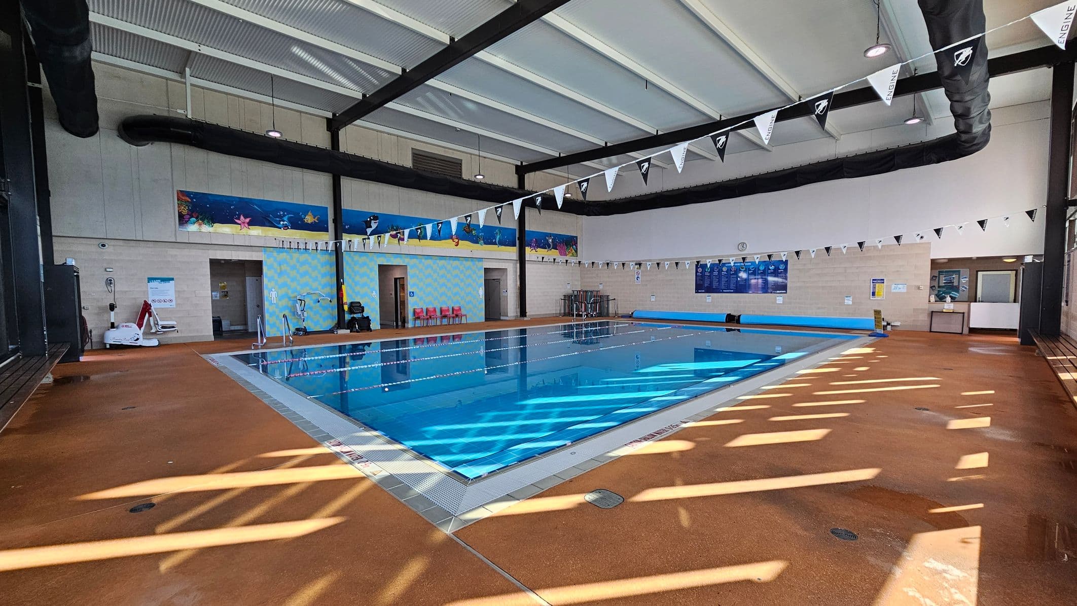 An indoor swimming pool with clear blue water, surrounded by a brown tiled deck. On the left, there are aquatic-themed murals on the walls and a few red chairs. The ceiling is high with exposed beams and large black ventilation ducts. Triangular flags are strung across the pool. On the right, a notice board and a small reception area are visible. The floor has a pattern of light and shadow from the ceiling.