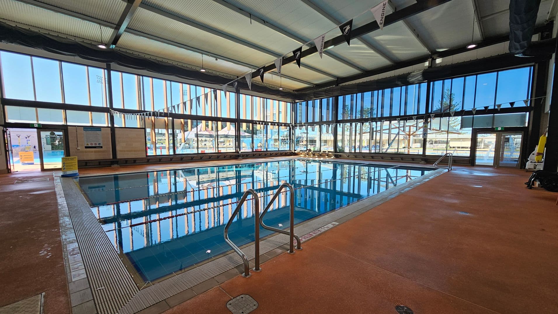 An indoor swimming pool with clear blue water is surrounded by large glass windows on three sides, allowing natural light to flood the space. The ceiling is high with exposed beams and lighting fixtures. The floor is a reddish-brown non-slip surface. Metal handrails lead into the pool, and there are safety signs and equipment visible. Outside, through the windows, there are glimpses of an outdoor area with structures and trees.
