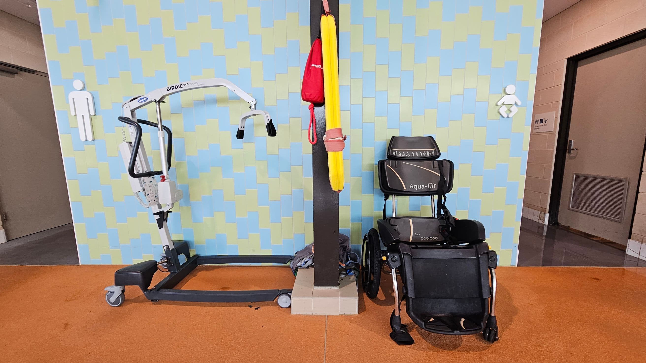 A room with a patterned wall in blue and green shades. On the left, there's a mobile hoist labelled "Birdie Evo Plus" with a male restroom sign above it. In the centre, a vertical pole holds a yellow flotation device and a red bag. On the right, an "Aqua-Tilt" wheelchair is positioned beneath a baby changing sign. The floor is orange, and a door is visible in the background on the right.