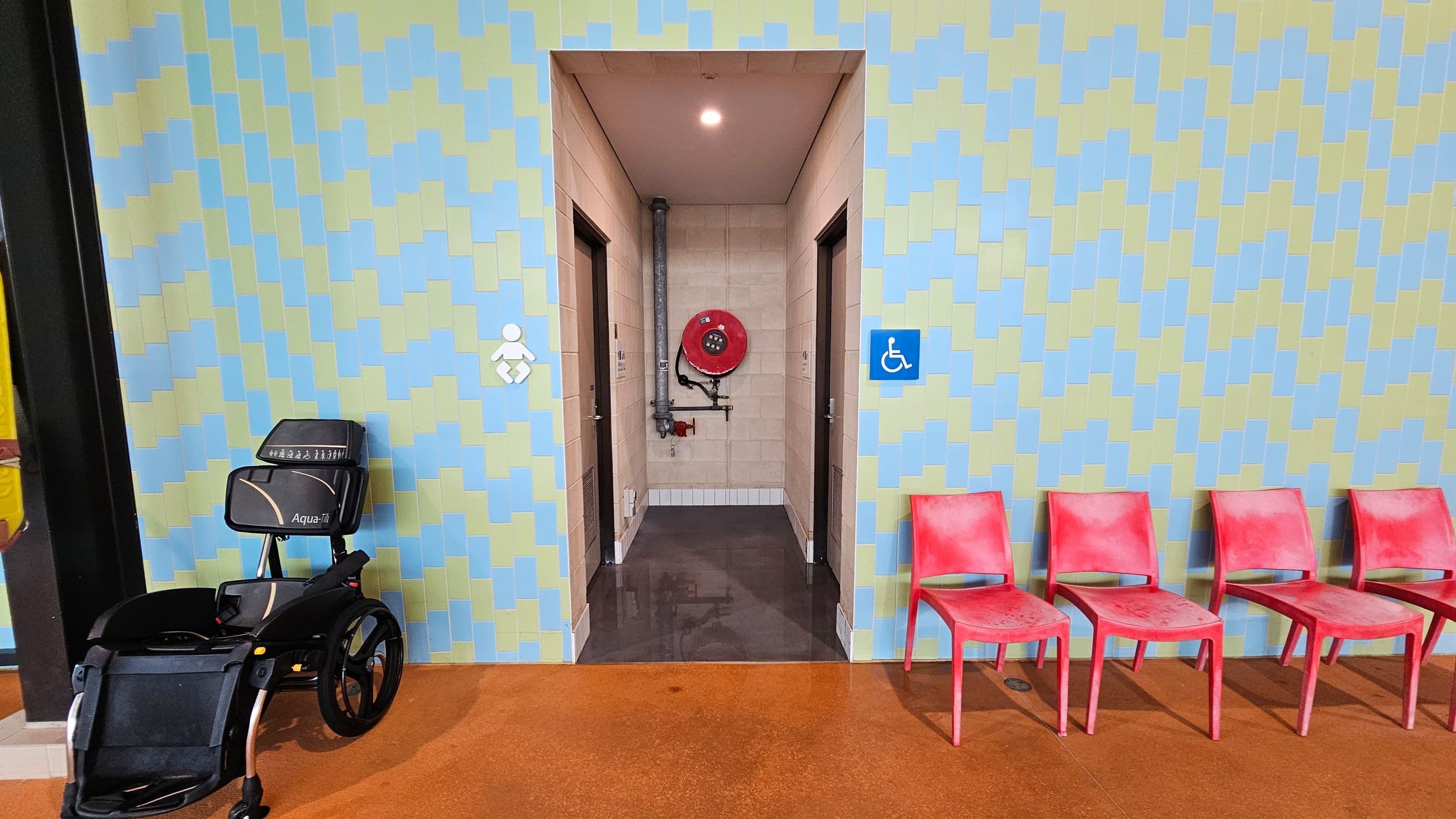A corridor with a patterned wall in blue and green tones. In the centre, there is an entrance with a fire hose reel mounted on the back wall. On the left, a black Aqua-Trek wheelchair is parked. On the right, four red plastic chairs are lined up against the wall. The floor is a reddish-brown colour, and there are symbols indicating accessibility and baby changing facilities near the entrance.