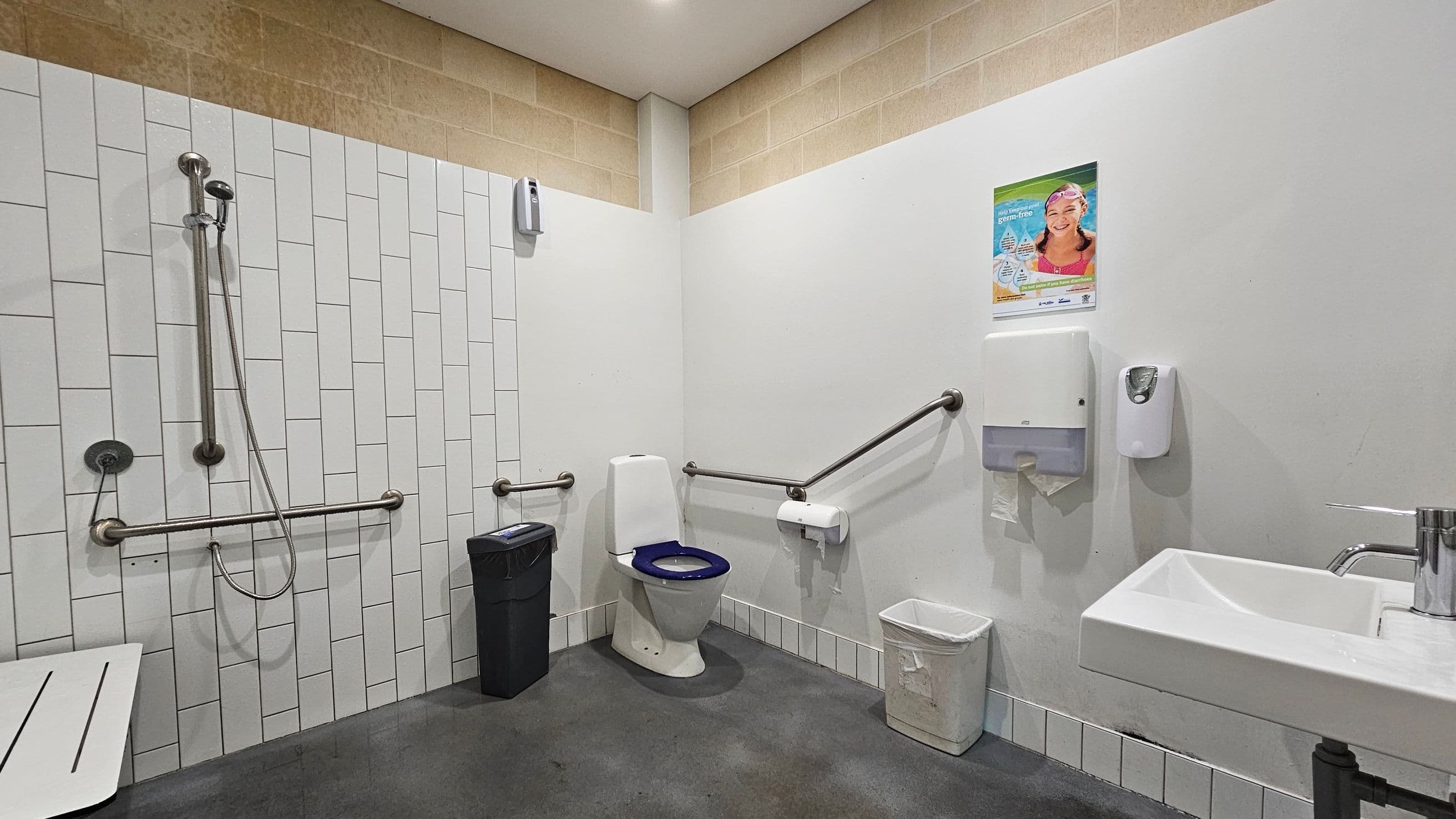 A public accessible bathroom with a shower on the left, featuring a handheld showerhead and grab bars. In the centre, there's a toilet with a blue seat, surrounded by grab bars. A black bin is next to the toilet, and a white bin is on the right. Above the bins, a poster and a paper towel dispenser are mounted on the wall. On the right, a white sink with a metal tap is visible. The floor is grey, and the walls are tiled with white and beige tiles.
