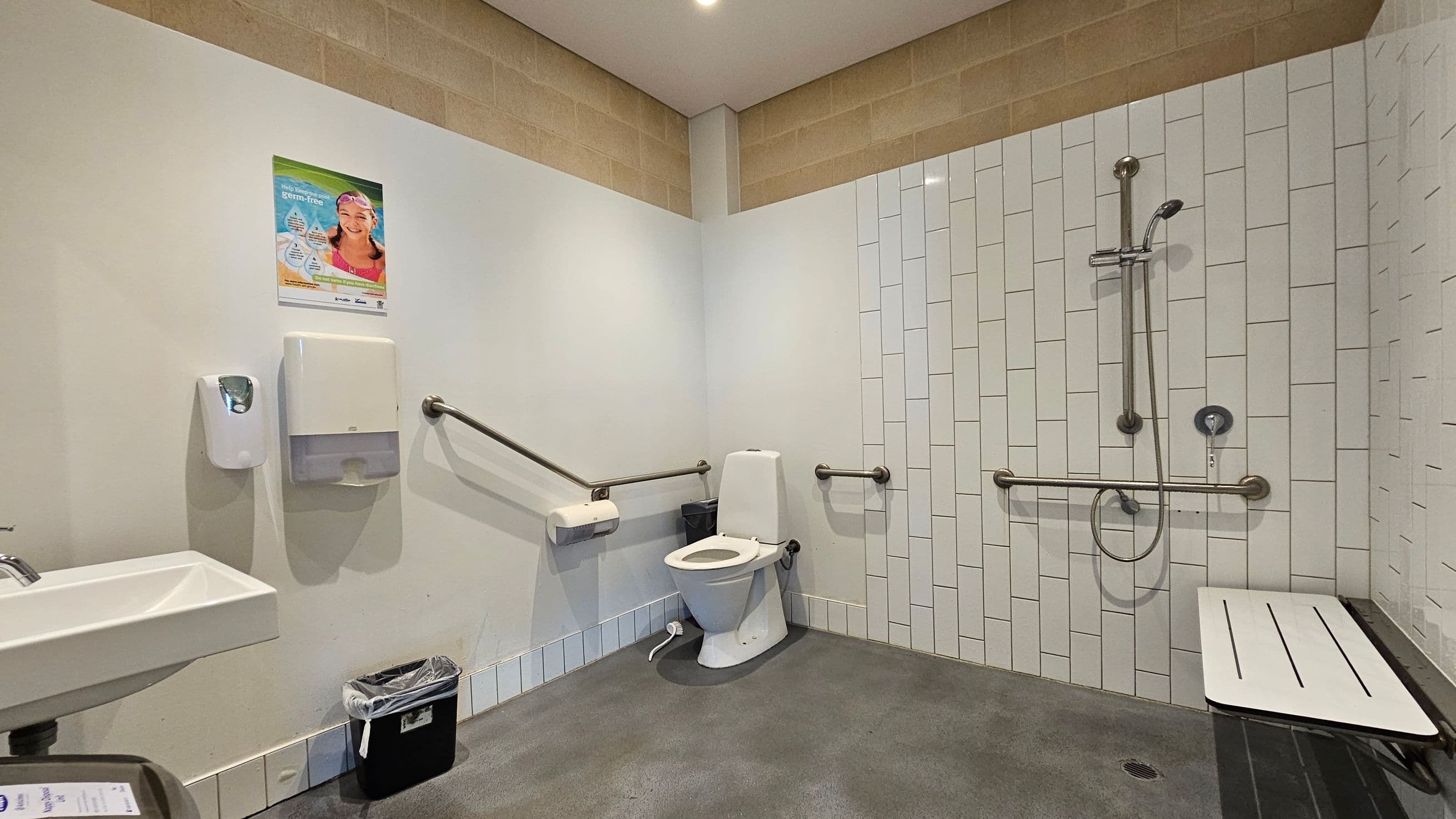 Accessible bathroom with a toilet in the centre, surrounded by grab bars. On the left, a sink with a soap dispenser and paper towel holder above a small bin. A poster is on the wall above. On the right, a shower area with a handheld showerhead and more grab bars, alongside a foldable shower seat. The walls are tiled, and the floor is non-slip. The ceiling is plain with recessed lighting.
