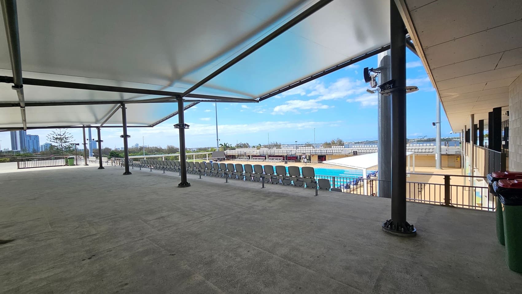 A covered outdoor area with a ceiling featuring recessed lights and a security camera. On the left, there's a door marked "Fire Hydrant" and "Fire Hose Reel." In the centre, a glass door leads to an indoor space with visible people. An elevator is adjacent to the door. On the right, a railing encloses a ramp or stairs, and several bins are visible. The background shows an open area with a canopy and some greenery.