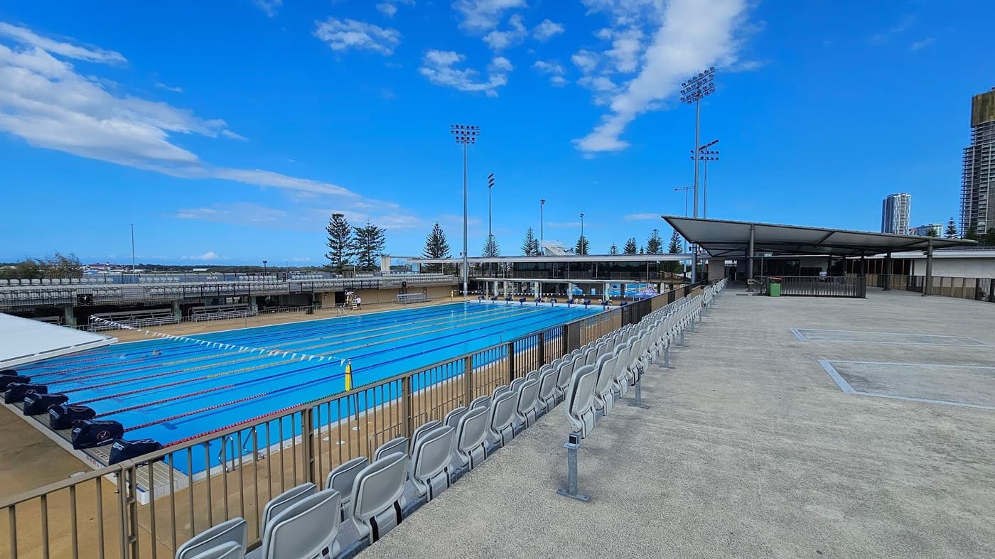 An outdoor swimming pool with multiple lanes is on the left, surrounded by a low fence. Empty spectator seating lines the poolside. On the right, a wide concrete area extends with more seating and a covered structure. Tall floodlights are visible in the background, along with a few trees and distant buildings.