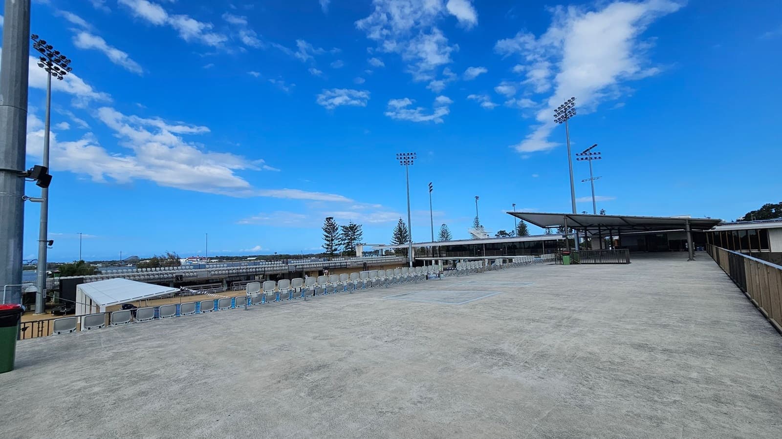 A large, open concrete area with rows of empty seats along the left side. Tall floodlights are visible in the background, along with some trees. On the right, there is a covered structure with a roof supported by metal beams. A railing runs along the right edge of the area. The sky is clear with a few clouds.