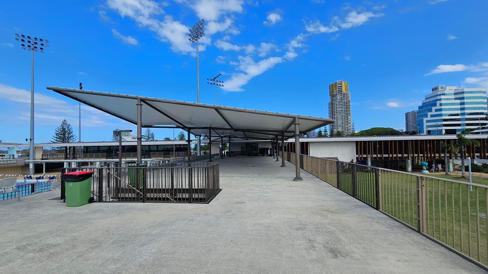 A concrete walkway leads to a covered area with a metal roof supported by poles. On the left, there is a fenced-off area with green bins and a swimming pool with lane dividers. Tall floodlights are visible in the background. On the right, a grassy area is bordered by a metal railing. In the distance, there are modern buildings, including a tall structure with yellow accents and a stepped white building.