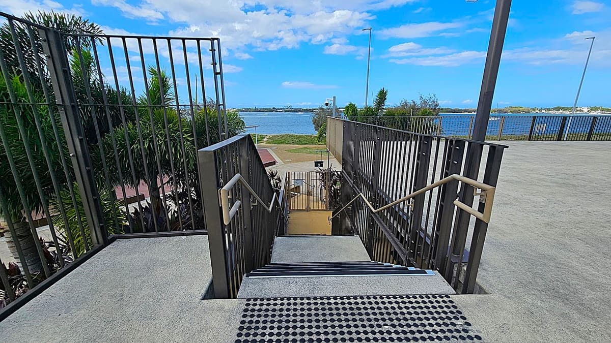 Concrete steps with metal railings descend towards a waterfront. On the left, lush green palm trees are visible, while the right side features a paved area with tall street lamps. In the background, a body of water stretches across the scene, with a distant shoreline and some buildings visible. The foreground shows a textured mat at the top of the stairs.