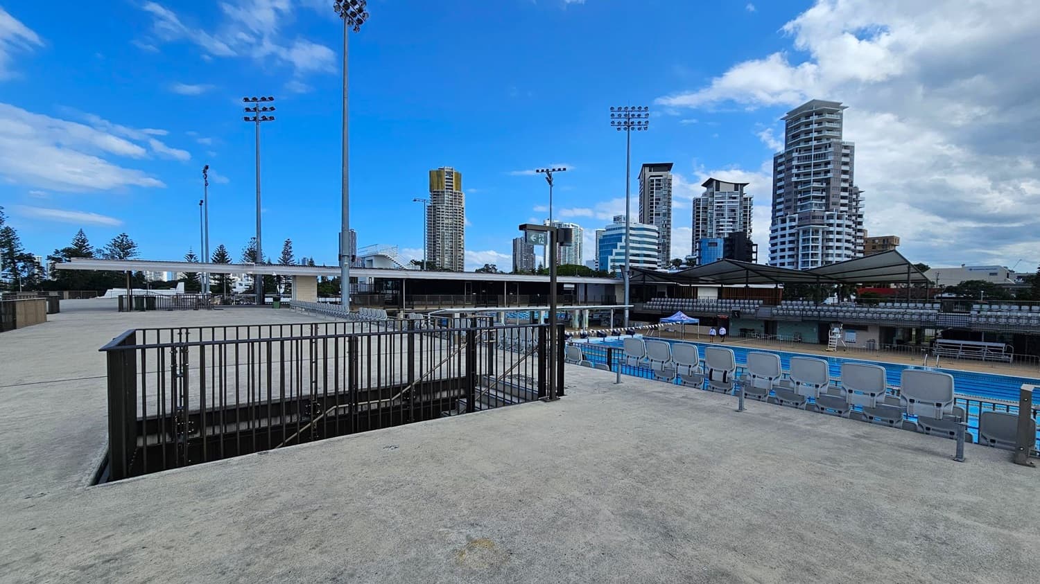 A concrete platform with a black metal railing on the left, leading to a staircase. In the middle, there are rows of blue stadium seats facing a swimming pool. Tall floodlights are visible, and in the background, there are several high-rise buildings. The area is open and spacious, with some trees visible in the distance.