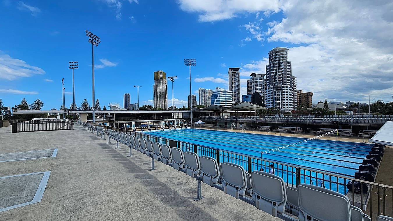 Outdoor swimming pool with multiple lanes on the right, surrounded by a fence. Empty tiered seating with grey chairs lines the left side. Tall floodlights are visible in the background, along with several high-rise buildings. The ground is paved, and there are some trees in the distance.