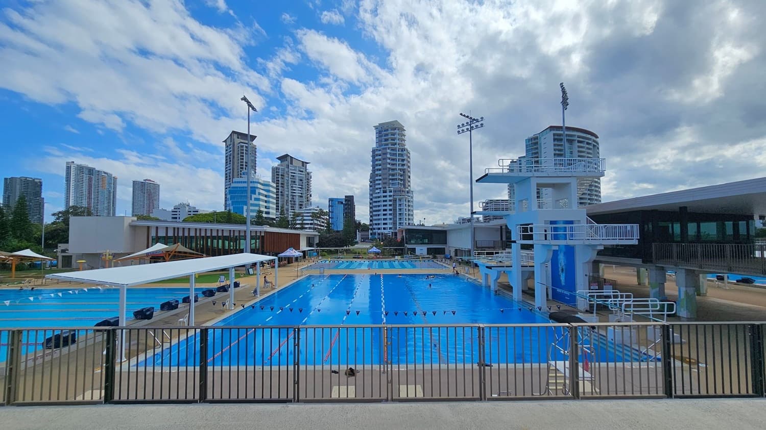 A large outdoor swimming pool with lanes is in the centre, surrounded by a metal fence in the foreground. On the right, there's a tall diving platform with multiple levels. To the left, there are shaded seating areas with white canopies. In the background, several high-rise buildings are visible against a partly cloudy sky. The pool area is bordered by a beige deck.