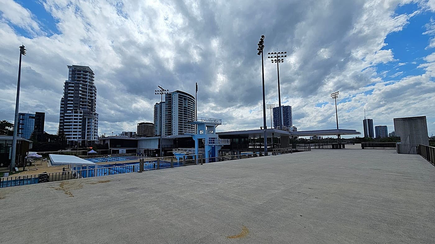 A large concrete platform in the foreground with a few patches of discolouration. In the middle, there is a swimming pool complex with diving platforms and a few people visible. Tall floodlights are positioned around the area. In the background, several modern high-rise buildings are visible against a cloudy sky. The scene is urban with a mix of sports and residential structures.