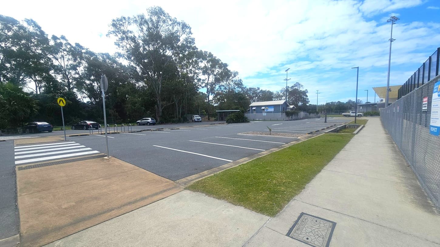 A car park with marked spaces is on the left, bordered by a row of trees and a few parked cars. A zebra crossing leads to a footpath. On the right, a concrete footpath runs alongside a chain-link fence with signs. A small building is visible in the background, and tall floodlights are positioned throughout the area. The sky is partly cloudy.