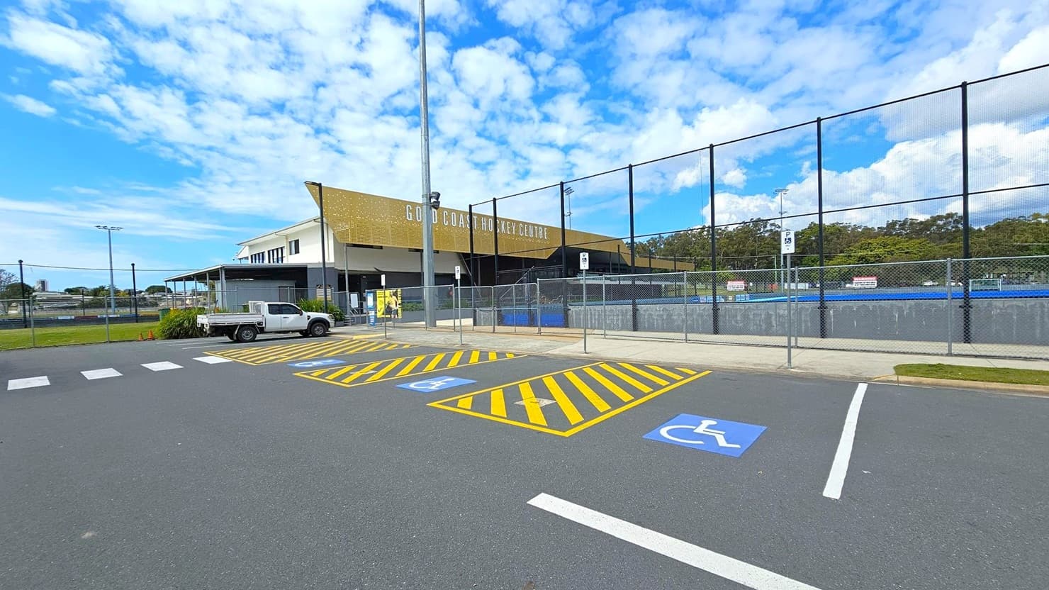 A sports facility with a sign reading "Gold Coast Hockey Centre" on the building. On the left, a white utility vehicle is parked near a pedestrian crossing. In the foreground, there are marked parking spaces for disabled access with blue and yellow lines. To the right, a fenced area encloses a blue sports field. The background features trees and a clear sky.