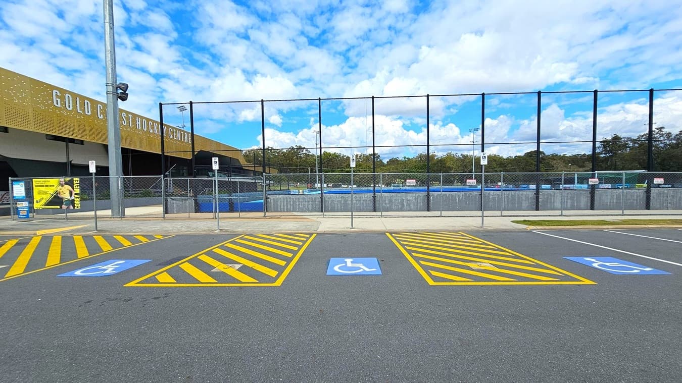 A parking area with designated disabled parking spaces marked by blue symbols on the asphalt. Yellow diagonal lines separate the spaces. In the background, a building with the sign "Gold Coast Hockey Centre" is visible, along with a tall fence enclosing a sports field. A large light pole stands to the left, and a poster is displayed on the fence. Trees are visible beyond the fence, under a partly cloudy sky.