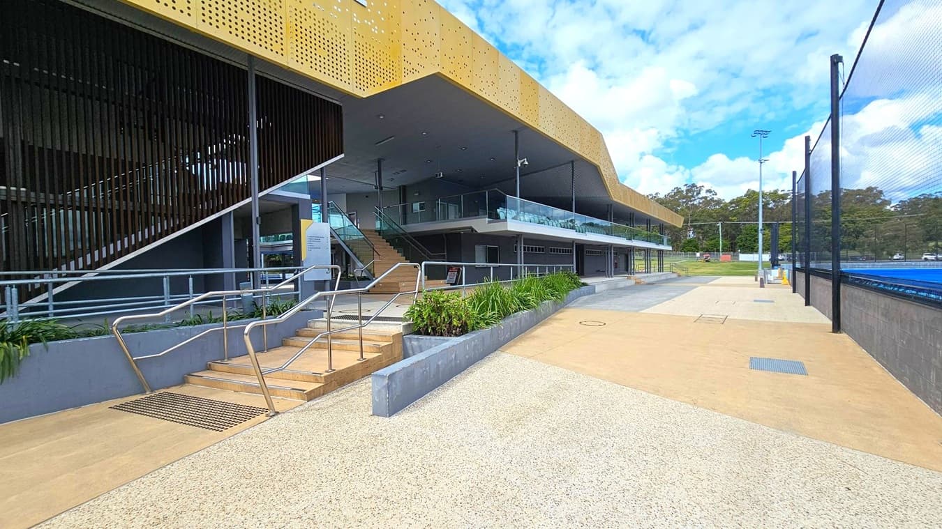 A modern building with a perforated gold-coloured facade on the upper level. On the left, there are stairs with metal railings leading to the entrance, flanked by a ramp and greenery. The middle section features a glass balcony and a covered walkway. On the right, a blue sports court is enclosed by a high fence. The ground is paved with a mix of light and dark concrete. In the background, there are trees and a few tall light poles.