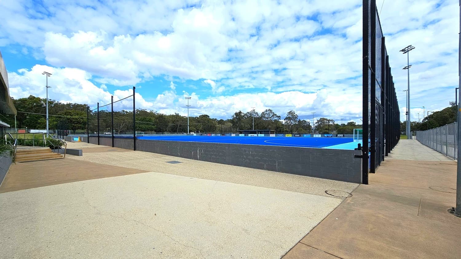 On the left, there are steps leading up to a building with a railing and some greenery. In the centre, a blue sports field is enclosed by a black mesh fence. The right side features a pathway running alongside the fence, with tall floodlights visible. In the background, there are trees and more floodlights surrounding the field. The ground is a mix of light and dark concrete.
