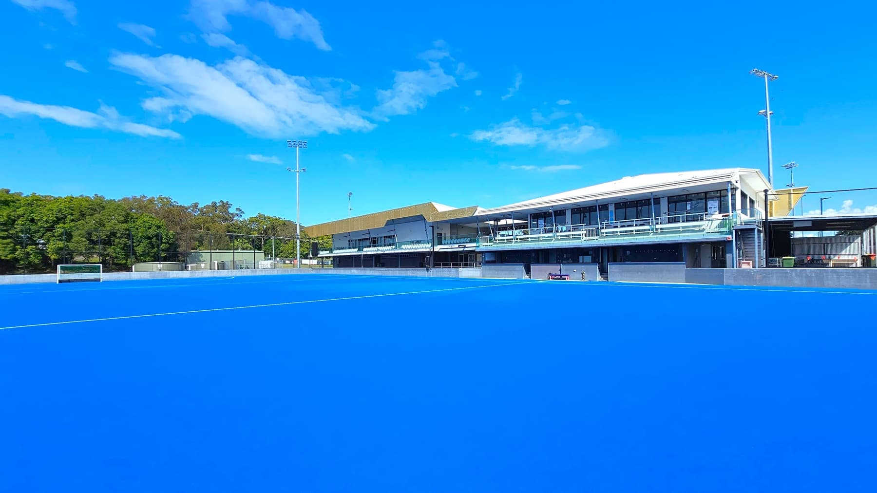 A bright blue hockey field dominates the foreground, with a goalpost visible on the left. In the background, there is a large building with a modern design, featuring multiple levels and glass railings. To the left, dense green trees form a natural border. Tall floodlights are positioned around the field. The sky is clear and blue.