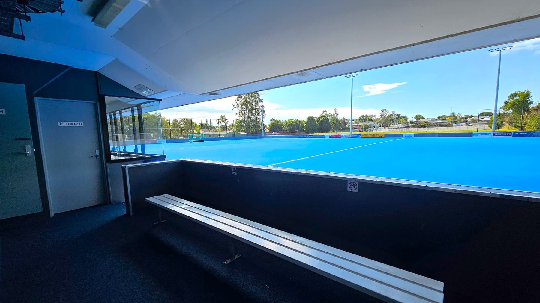 View from inside a sports facility overlooking a bright blue hockey field. In the foreground, a bench with metal slats is visible. To the left, a door marked "Tech Bench" is seen. The background shows the field with white lines, surrounded by trees and a few buildings. The ceiling is white with some structural elements visible.
