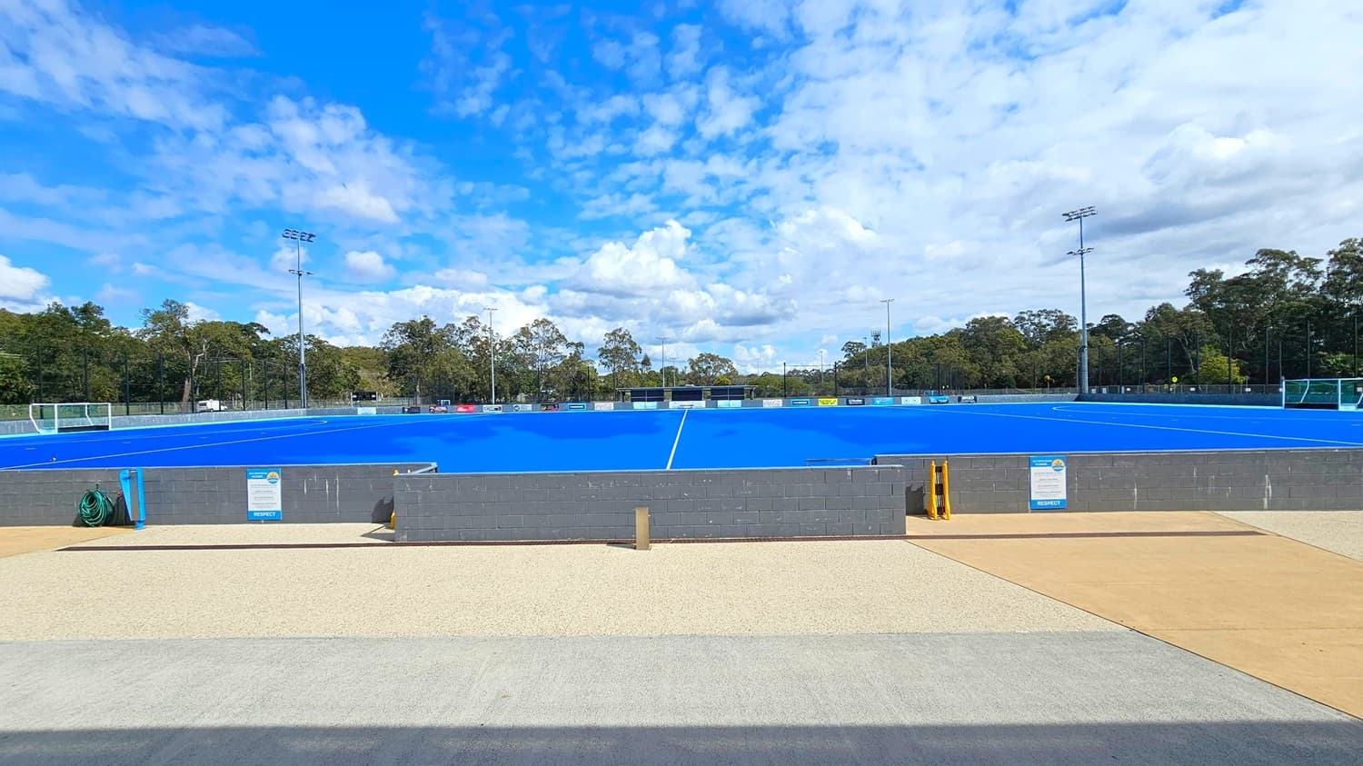 A blue hockey field is in the centre, surrounded by a low grey wall. Two goalposts are visible at each end of the field. Tall floodlights are positioned around the perimeter. In the background, there are trees lining the edge of the field. The foreground features a beige and grey paved area with a green hose coiled on the left side. Signs are attached to the wall, and a yellow barrier is visible near the centre.