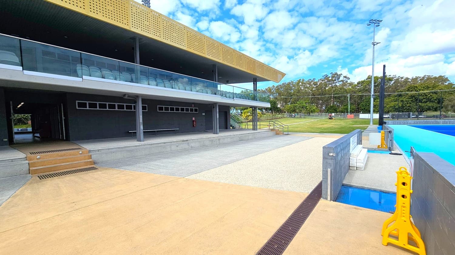 A sports facility with a two-level structure on the left, featuring a perforated yellow facade and glass railings. The lower level has a dark wall with a bench and a fire extinguisher. In the foreground, there is a concrete area with a ramp and steps. On the right, a blue sports field is visible, bordered by a low wall with yellow barriers. In the background, there are grassy areas and trees, with tall floodlights towering above.
