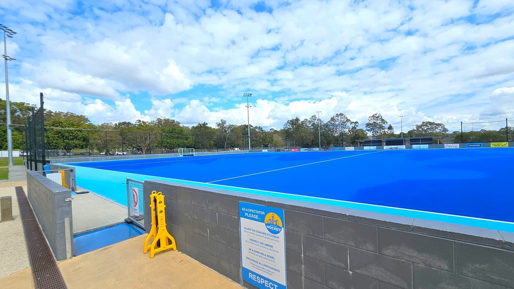 A bright blue hockey field is in the centre, surrounded by a low grey wall. On the left, a tall metal fence and a yellow caution sign are visible. In the background, there are trees and several floodlights. A sign on the wall in the foreground lists spectator guidelines. The ground is a mix of concrete and gravel.