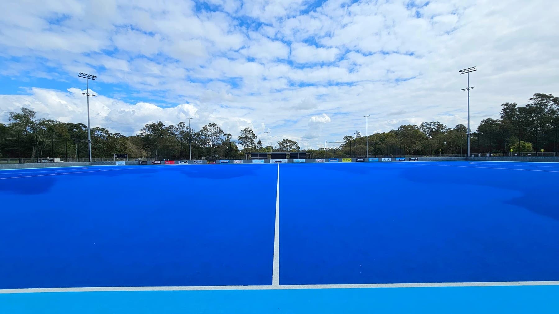 A blue sports field with a white centre line running vertically. Surrounding the field are tall floodlights and a perimeter fence with various advertisements. In the background, there are trees lining the edge of the field. The sky is filled with clouds.