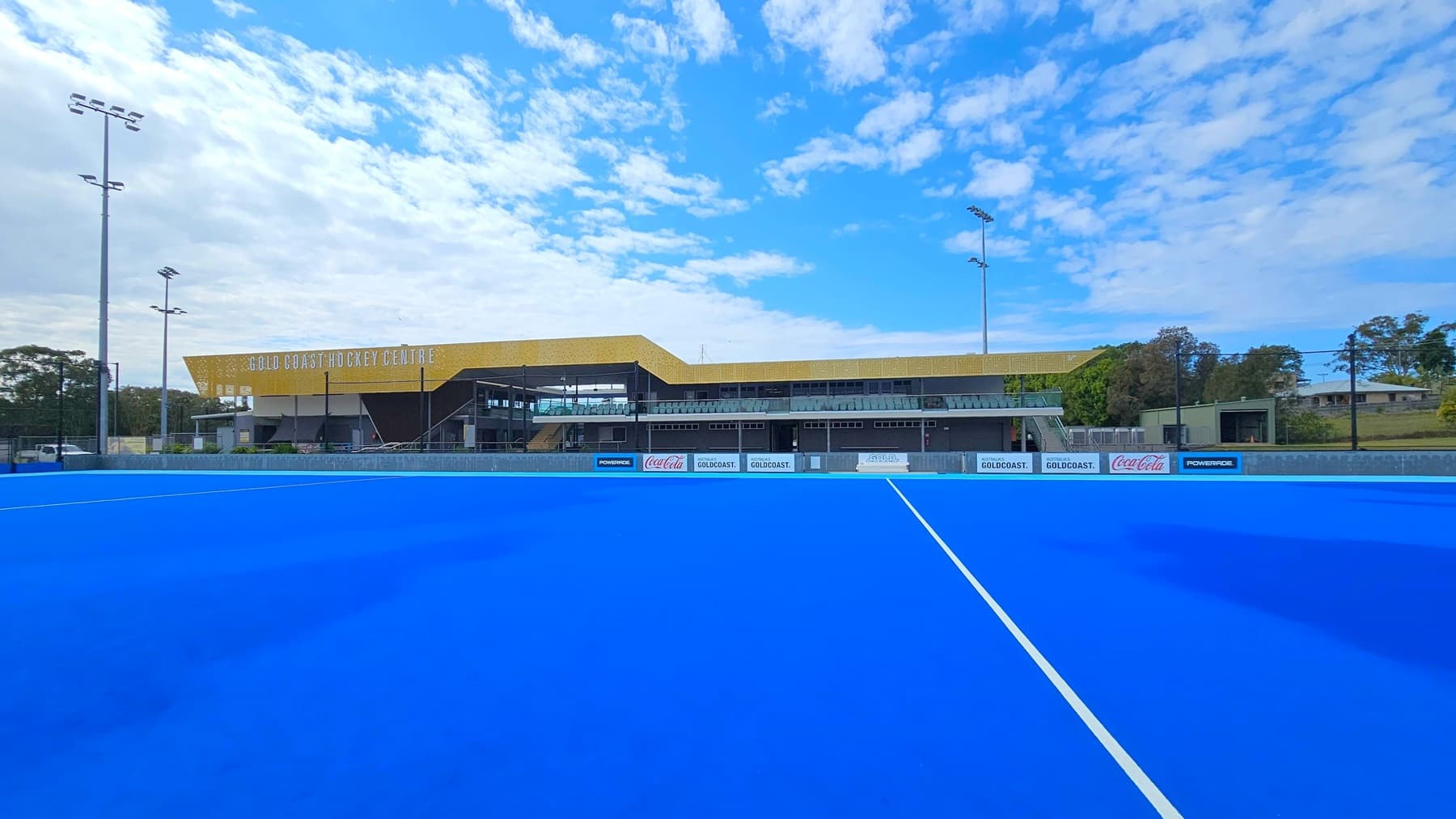 A bright blue hockey field stretches across the foreground, with a white line running through it. In the background, the Gold Coast Hockey Centre building is visible, featuring a yellow roof and a row of seating. Tall floodlights are positioned on both sides of the field. Trees and a few small structures are visible to the right, while the left side shows more trees and a parked vehicle. The sky is partly cloudy.