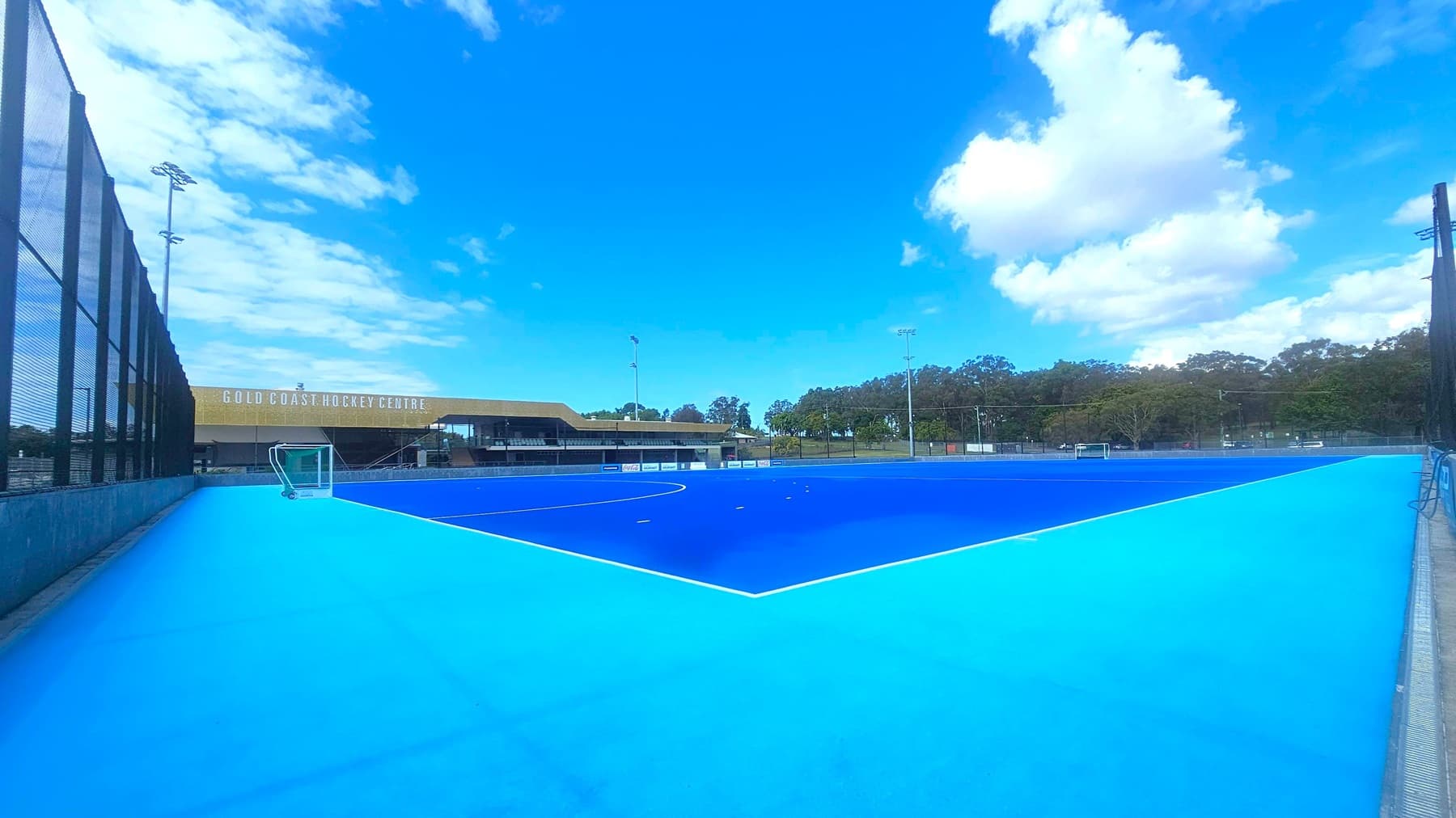 A blue hockey field with a lighter blue border. On the left, a tall black fence runs alongside the field. In the background, a building with the sign "Gold Coast Hockey Centre" is visible. Trees line the horizon, and there are several tall floodlights around the field. A small goal is positioned near the centre of the image. The sky is mostly clear with a few clouds.