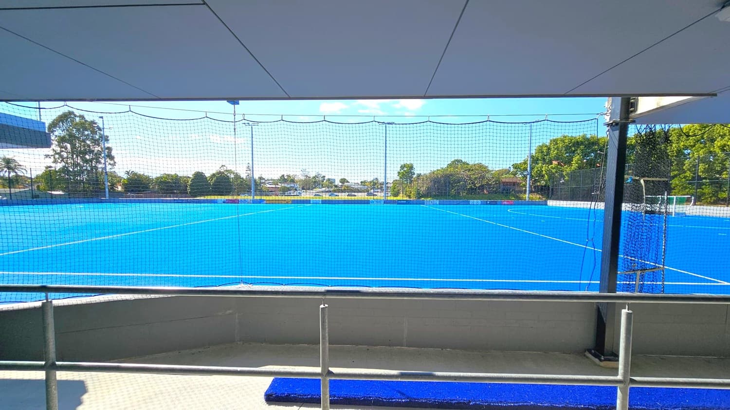 A bright blue artificial turf sports field is visible through a netted barrier. In the foreground, there is a railing and a small section of blue matting on a grey floor. The background features trees and some buildings, with a clear view of the horizon. The ceiling is a flat, grey structure with panels.