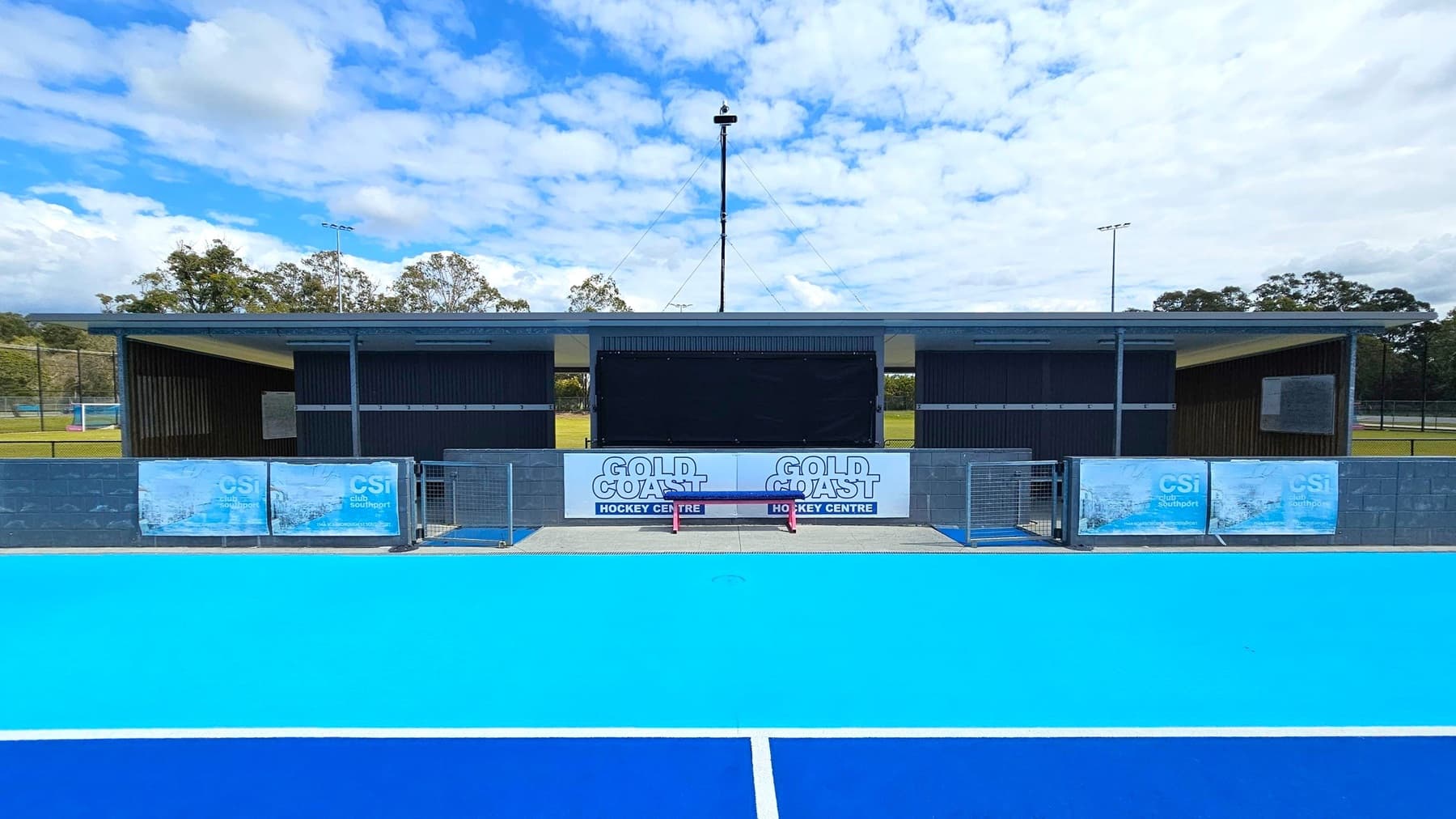 A sports facility with a bright blue hockey field in the foreground. In the middle, there is a building with a dark exterior and a central scoreboard. Signs reading "Gold Coast Hockey Centre" are displayed on the building. The background features trees and floodlights. The ground is a vivid blue, and the building is flanked by banners with "CSI Club Southport" on a low wall.