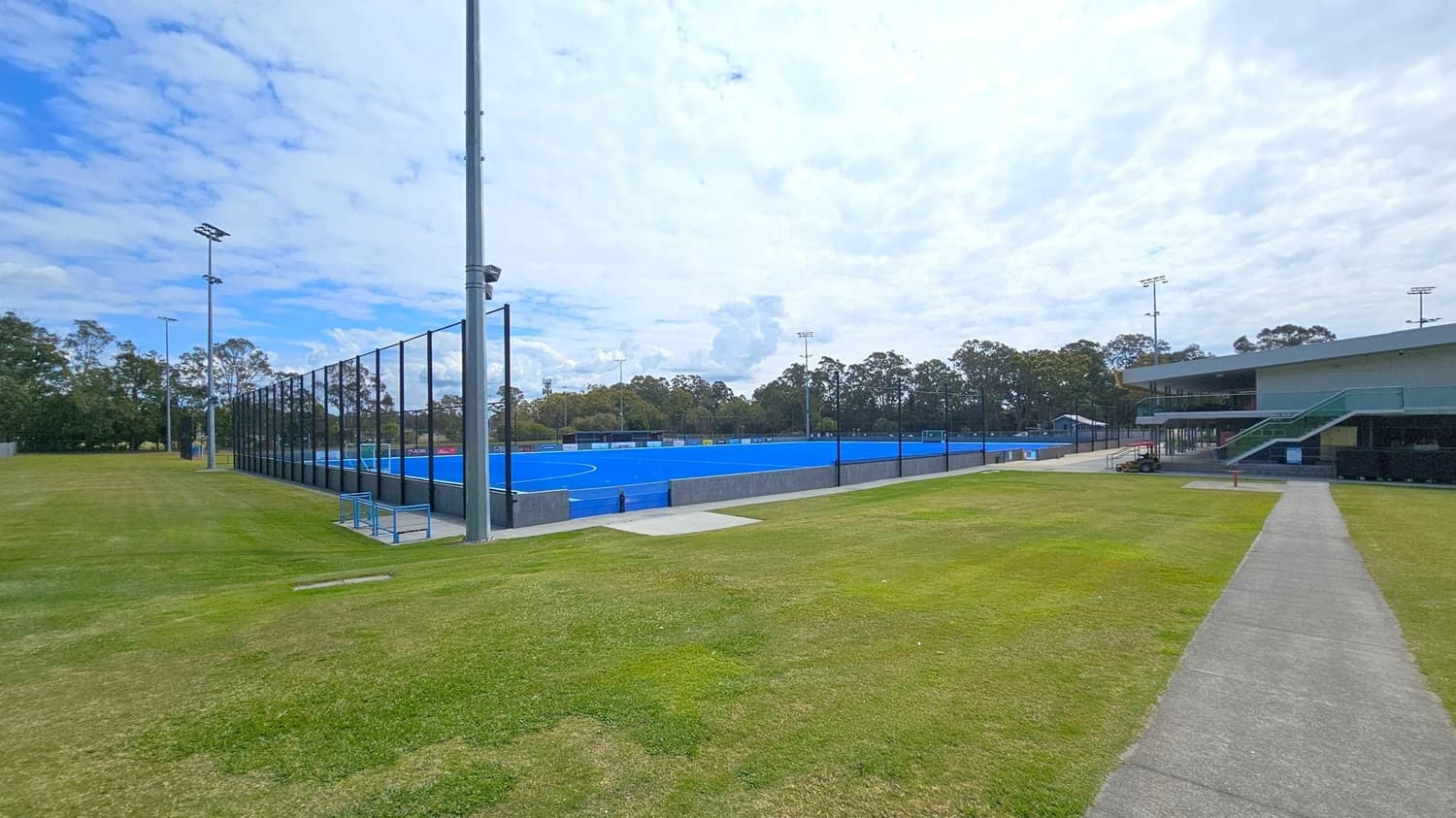 A sports facility with a blue artificial turf field in the centre, surrounded by a tall black fence. On the left, there is a grassy area with tall floodlights. On the right, a modern building with a staircase leading to an upper level. A concrete path runs from the foreground towards the building. Trees line the background, providing a natural border to the facility.