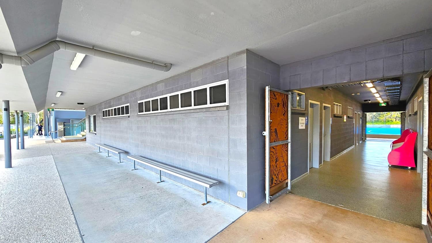 A covered outdoor area with a concrete floor and a grey ceiling featuring fluorescent lights. On the left, there are metal benches along a wall with small rectangular windows. The area leads to a corridor on the right, with doors and a bright pink chair visible inside. The corridor has a mix of brown and green flooring. In the background, there are steps leading to a blue structure, and a person is visible near the entrance.