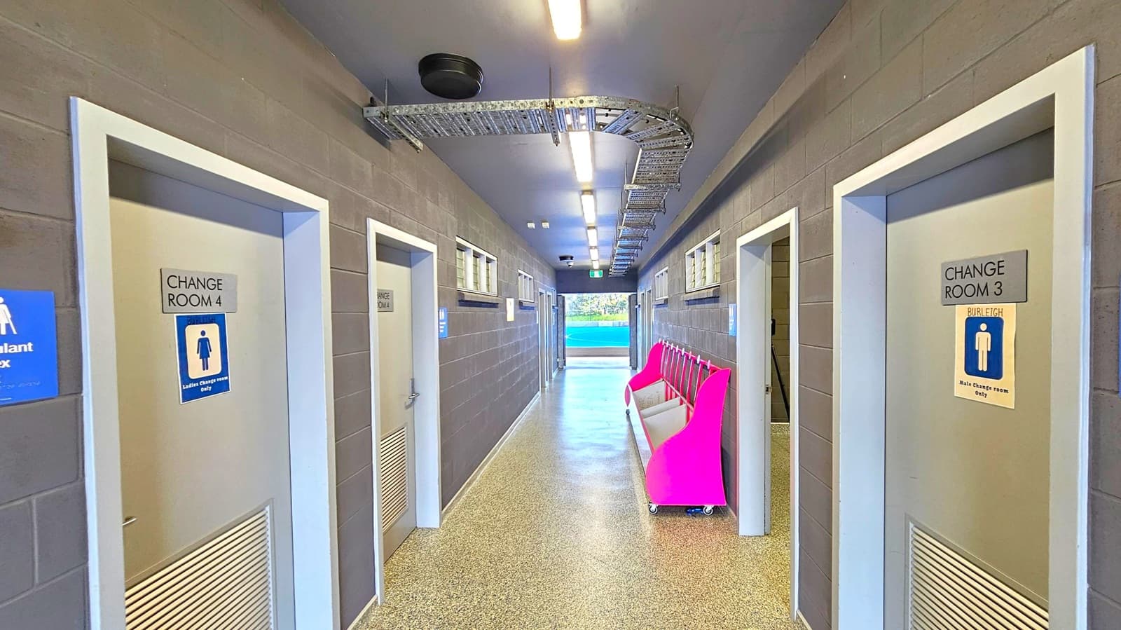 A corridor with grey brick walls and a speckled floor. On the left, doors labelled "Change Room 4" and "Change Room 1" are visible, with a sign for a defibrillator box. On the right, doors labelled "Change Room 3" and "Change Room 2" are present. A bright pink bench with wheels is positioned against the right wall. The ceiling has exposed metal cable trays and fluorescent lights. At the end of the corridor, there is an open doorway leading to an outdoor area with a blue surface.