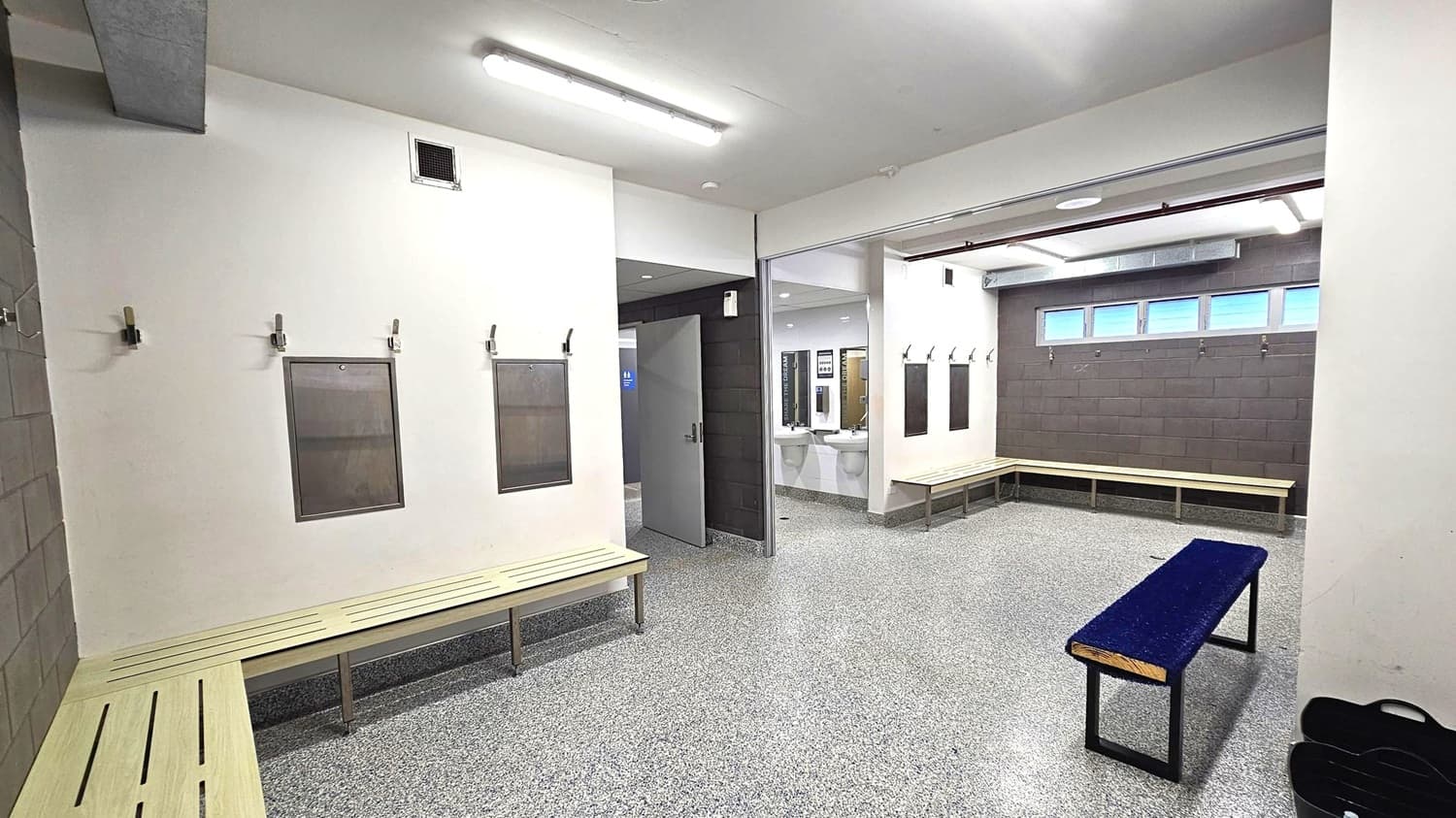 A locker room with grey speckled flooring and white walls. On the left, there are wooden benches with hooks and notice boards above them. In the centre, an open doorway leads to a washroom area with sinks and mirrors. On the right, more benches line the walls, with small windows above. The ceiling has fluorescent lights and exposed ductwork. A dark bench is positioned in the foreground.