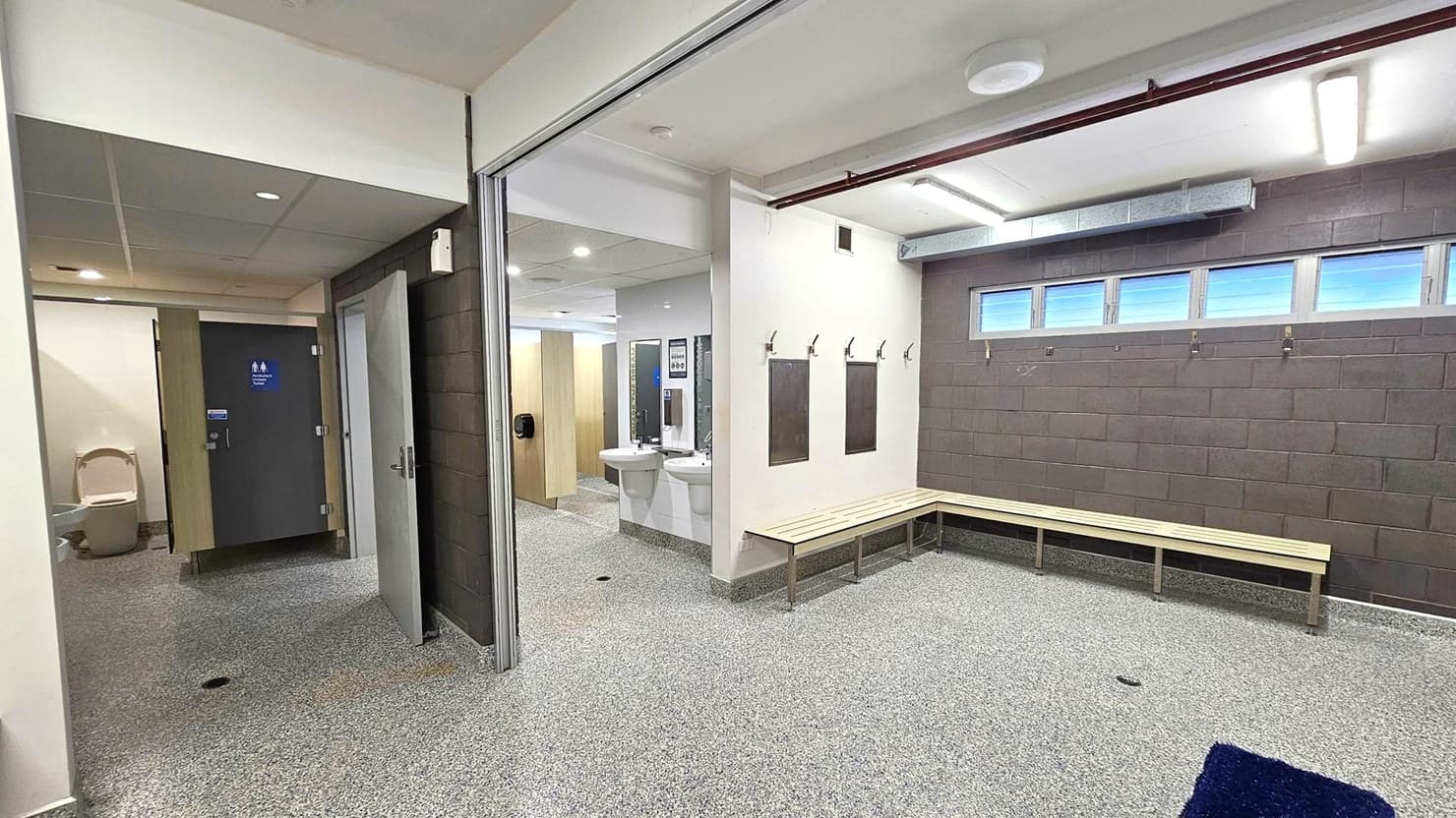 A changing room with a speckled grey floor and white ceiling. On the right, there is a long wooden bench against a dark brick wall with small windows above. Several hooks are mounted on the wall. In the centre, two white sinks with mirrors are visible. On the left, there are toilet cubicles with wooden doors, one slightly open, revealing a toilet. The ceiling has recessed lighting, and a purple mat is on the floor near the bench.
