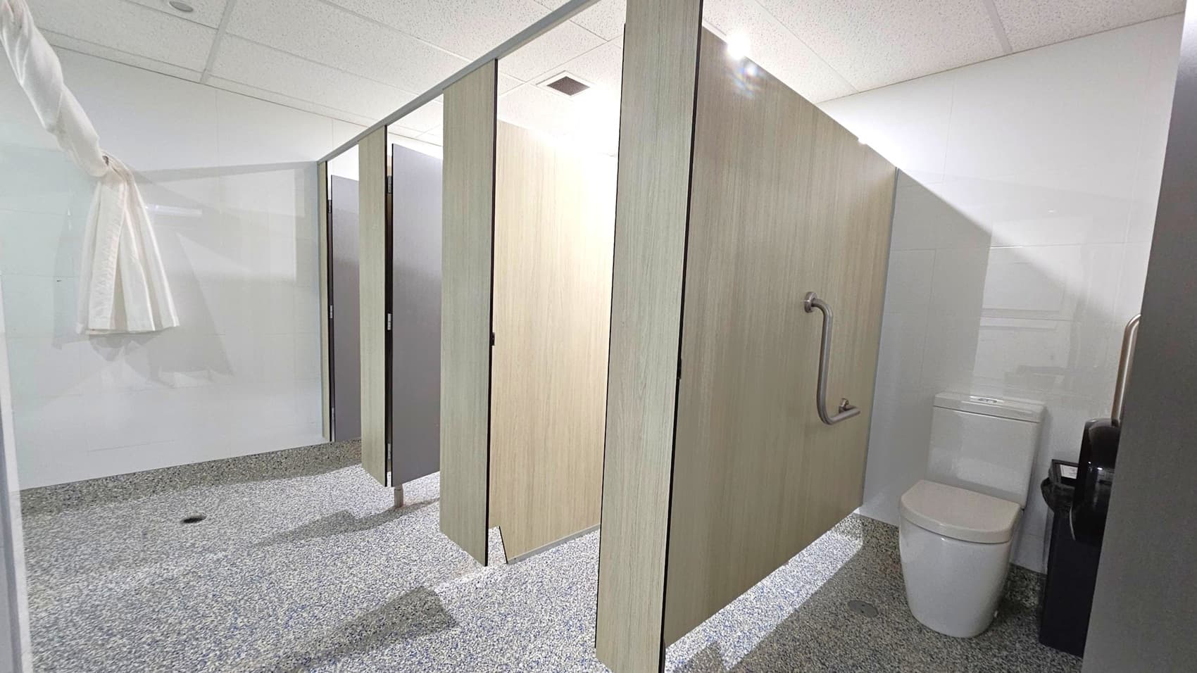 A public restroom with multiple stalls. On the right, a toilet is visible in a stall with a green door and a metal grab bar. The floor is speckled grey, and the ceiling is white with recessed lighting. On the left, a white curtain is partially visible. The partitions between stalls are a mix of green and beige. The walls are white, and there is a black waste bin next to the toilet.