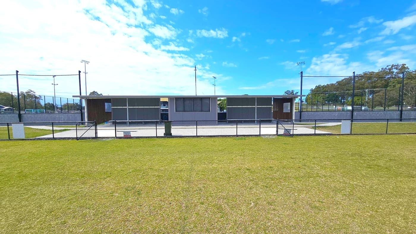 A sports facility with a low, rectangular building in the centre, featuring a flat roof and dark exterior. In front of the building is a concrete area bordered by a black metal fence. The foreground has a grassy field with patches of brown. On either side of the building, there are tall light poles and netted fencing. Trees are visible in the background on the right. The sky is partly cloudy with blue patches.