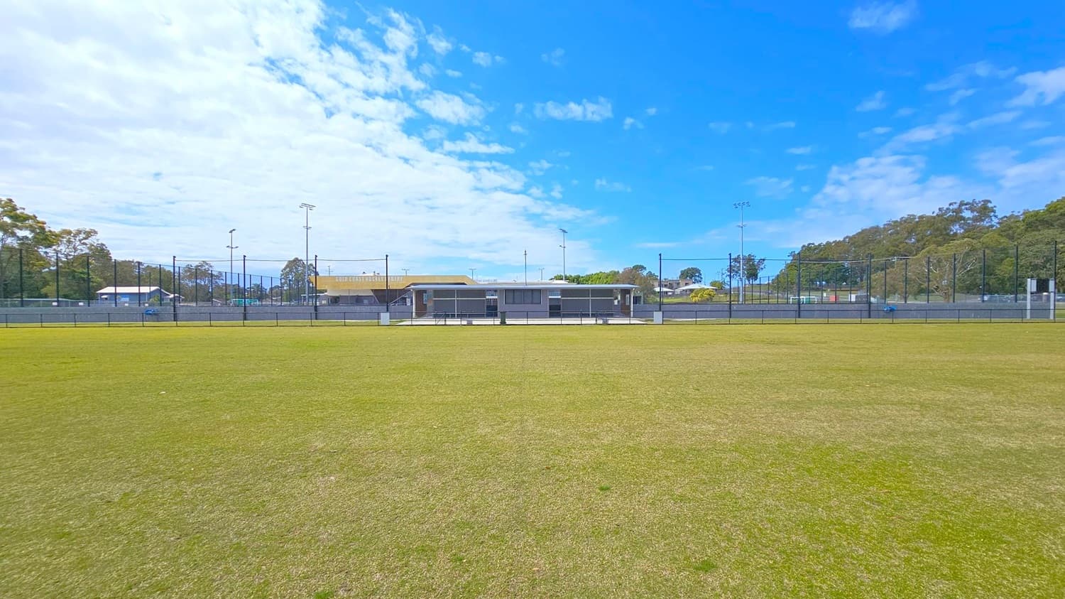 A grassy sports field with a slightly patchy surface in the foreground. In the middle, there is a fenced area with a low building, possibly a clubhouse or sports facility, featuring a covered seating area. The background has tall light poles and trees lining the perimeter. The sky is partly cloudy with patches of blue visible.