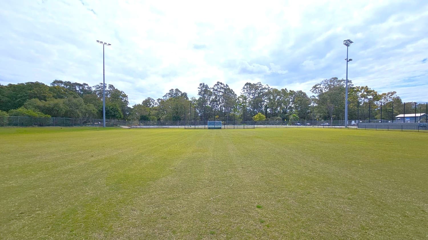 A grassy sports field with a goal in the centre background, surrounded by a fence. Tall floodlights are positioned on the left and right sides. Behind the fence, there are trees and some buildings visible. The grass has patches of wear, and the area is open and spacious.