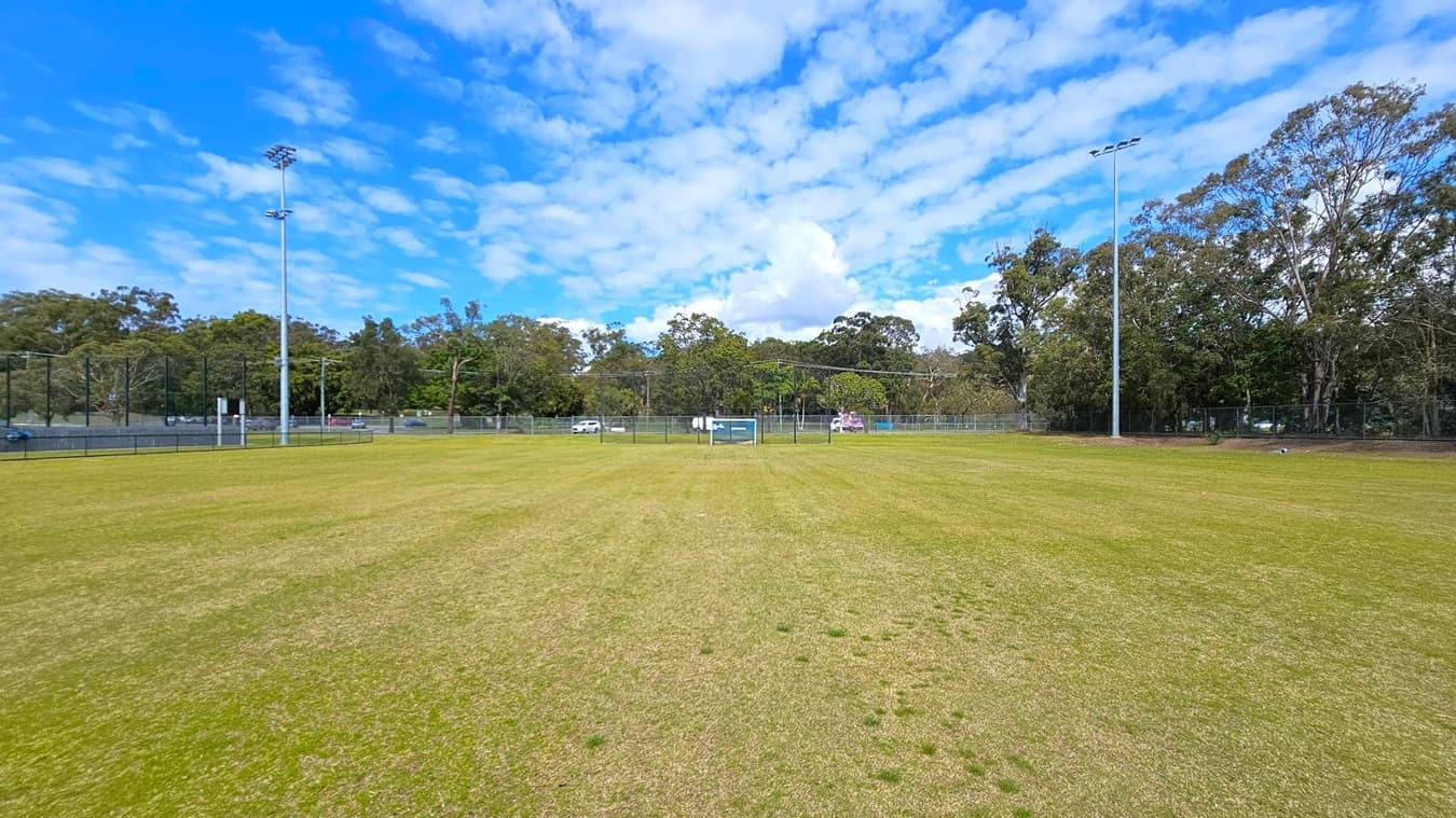 A grassy sports field with patches of brown and green grass. Two tall floodlights are positioned on the left and right sides. In the background, there is a line of trees and a fence, with a few vehicles visible behind the fence. The sky is partly cloudy.