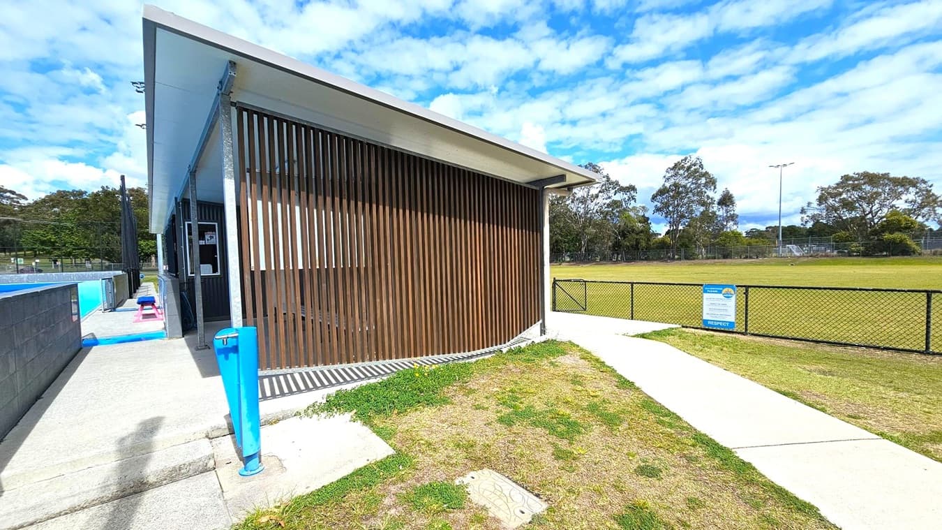 On the left, a building with vertical wooden slats and a blue water fountain is adjacent to a concrete path. A blue bench and a pool are visible further along the path. On the right, a grassy field is enclosed by a black chain-link fence with a sign attached. Trees and a sports field with goalposts are in the background. The ground is a mix of grass and concrete, and the building has a flat roof.