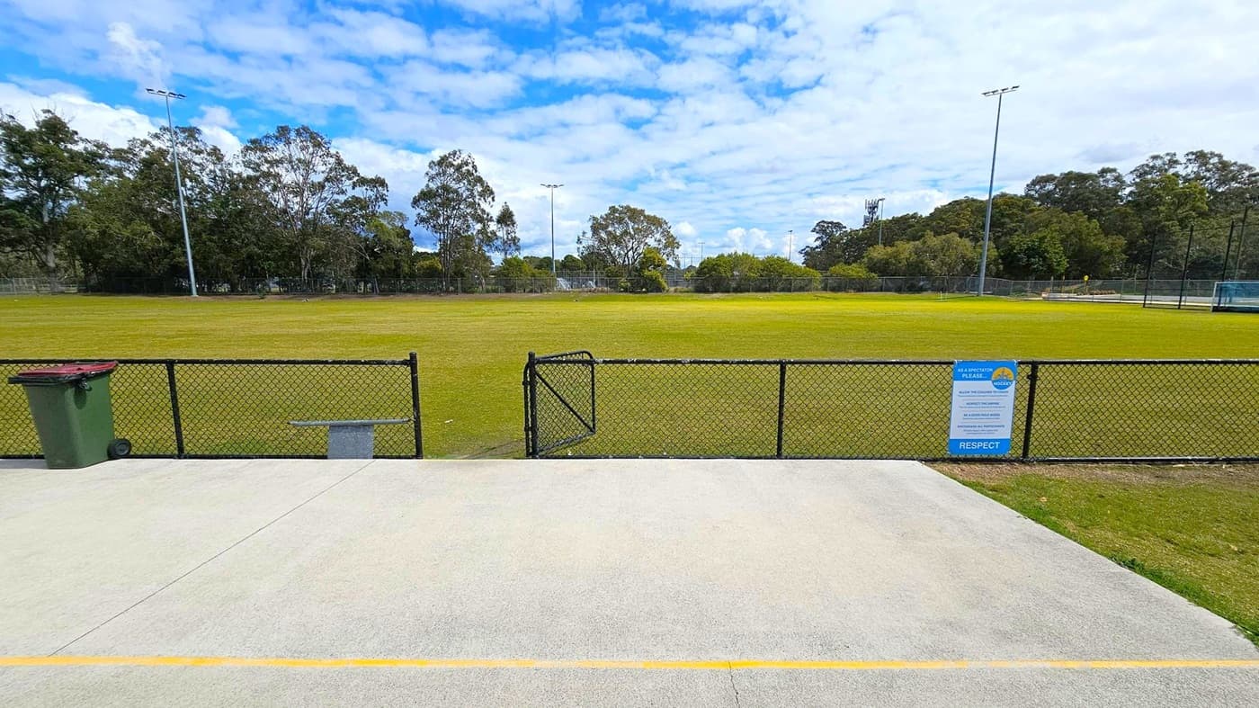 A grassy sports field is surrounded by a black chain-link fence with a gate in the centre. On the left, there's a green rubbish bin next to the fence. On the right, a sign is attached to the fence. Tall floodlights are visible around the field, and trees line the background. The foreground features a concrete path with a yellow line near the edge.