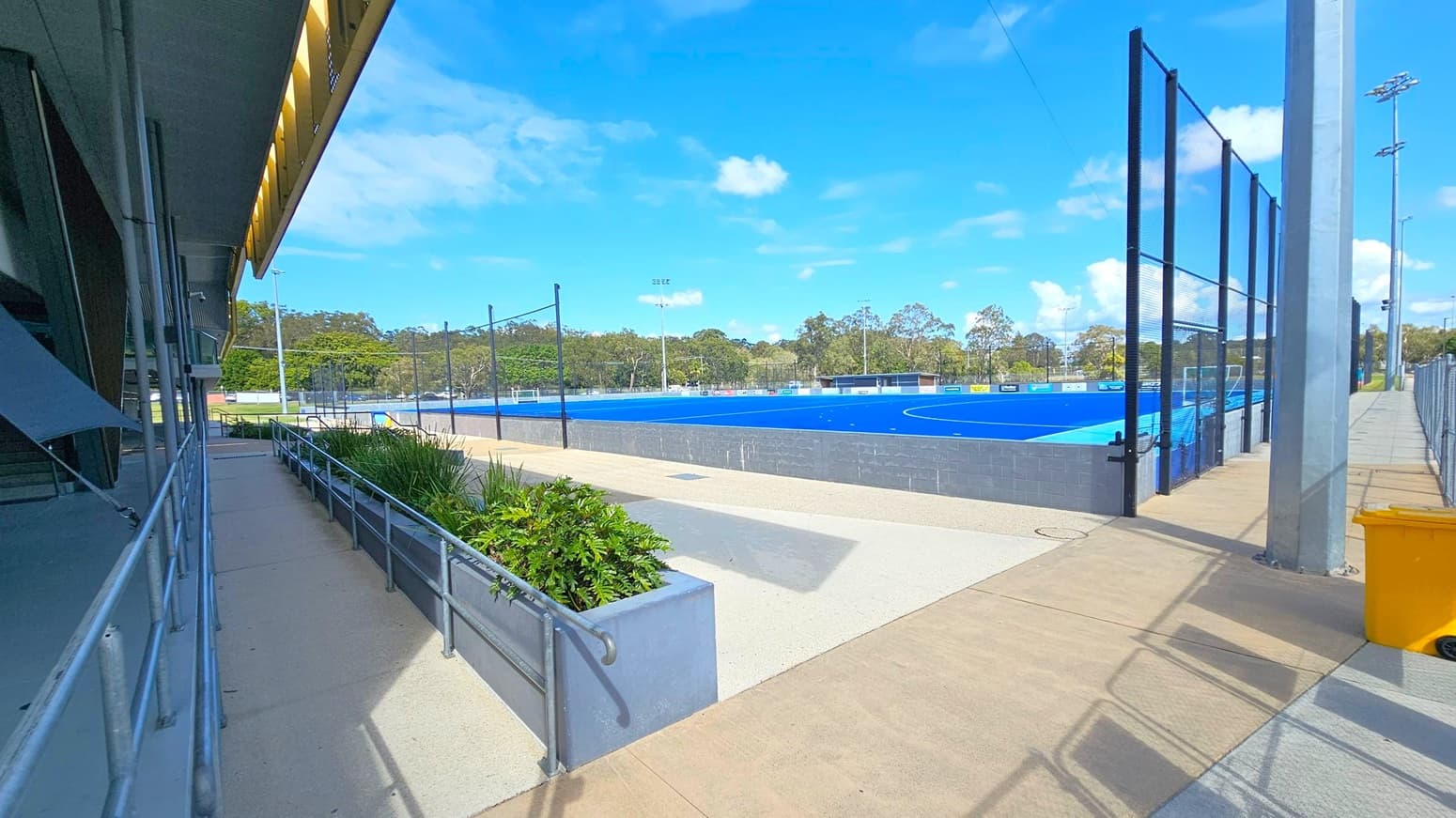 On the left, a building with a slanted roof and metal railings lines a concrete walkway. In the middle, a blue sports field is surrounded by a low grey wall and black mesh fencing. On the right, a tall metal pole and a yellow bin are near the entrance to the field. Trees and floodlights are visible in the background. The ground is a mix of concrete and paved surfaces.