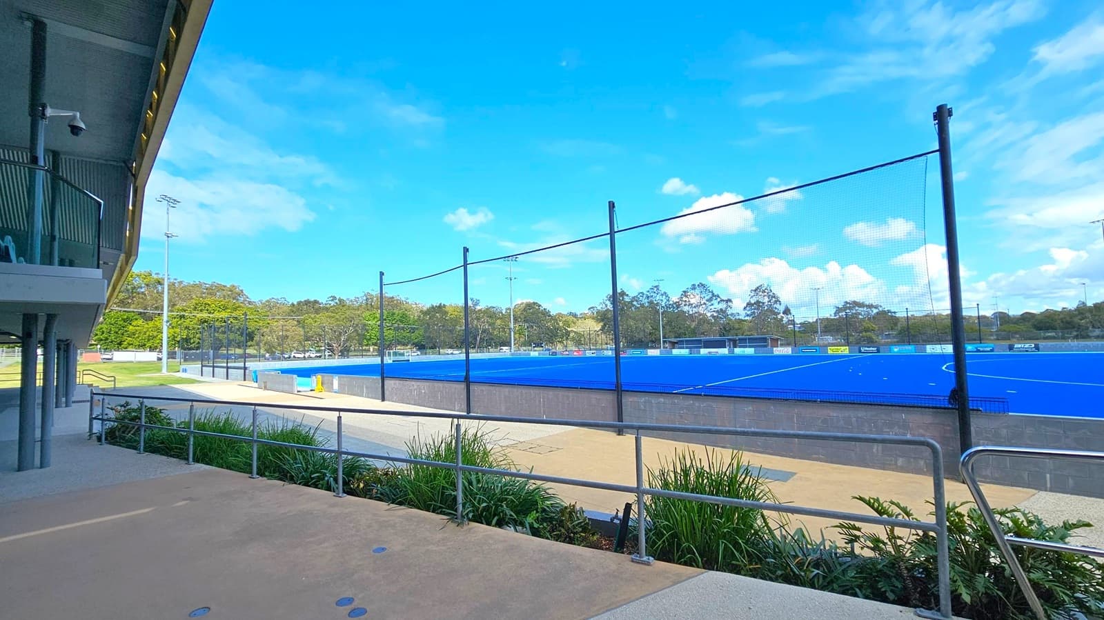 On the left, a building with a balcony and security camera is visible. The foreground has a concrete path with metal railings and green plants. To the right, a blue sports field is enclosed by a black mesh fence. In the background, there are trees and a few light poles. The sky is clear with some clouds.