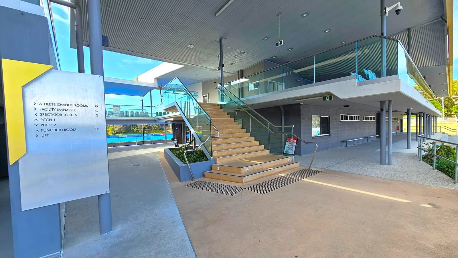 A modern building interior with a staircase in the centre leading to an upper level with glass railings. On the left, a sign lists facilities such as athlete change rooms, spectator toilets, and a function room. The floor is a smooth concrete surface, and the ceiling is lined with panels and lights. To the right, there is a corridor with windows and benches. In the background, a sports field is visible through large windows.