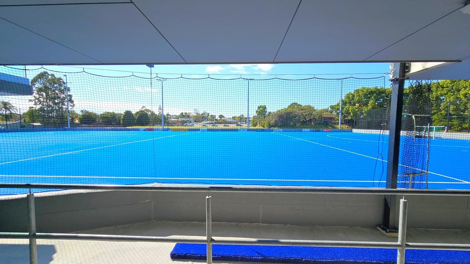 A bright blue sports field is visible through a netted barrier. In the foreground, there is a railing and a small section of blue matting on the ground. The background features trees and some buildings, with a clear view of the horizon. The ceiling is a flat, grey structure, providing shade over the viewing area.
